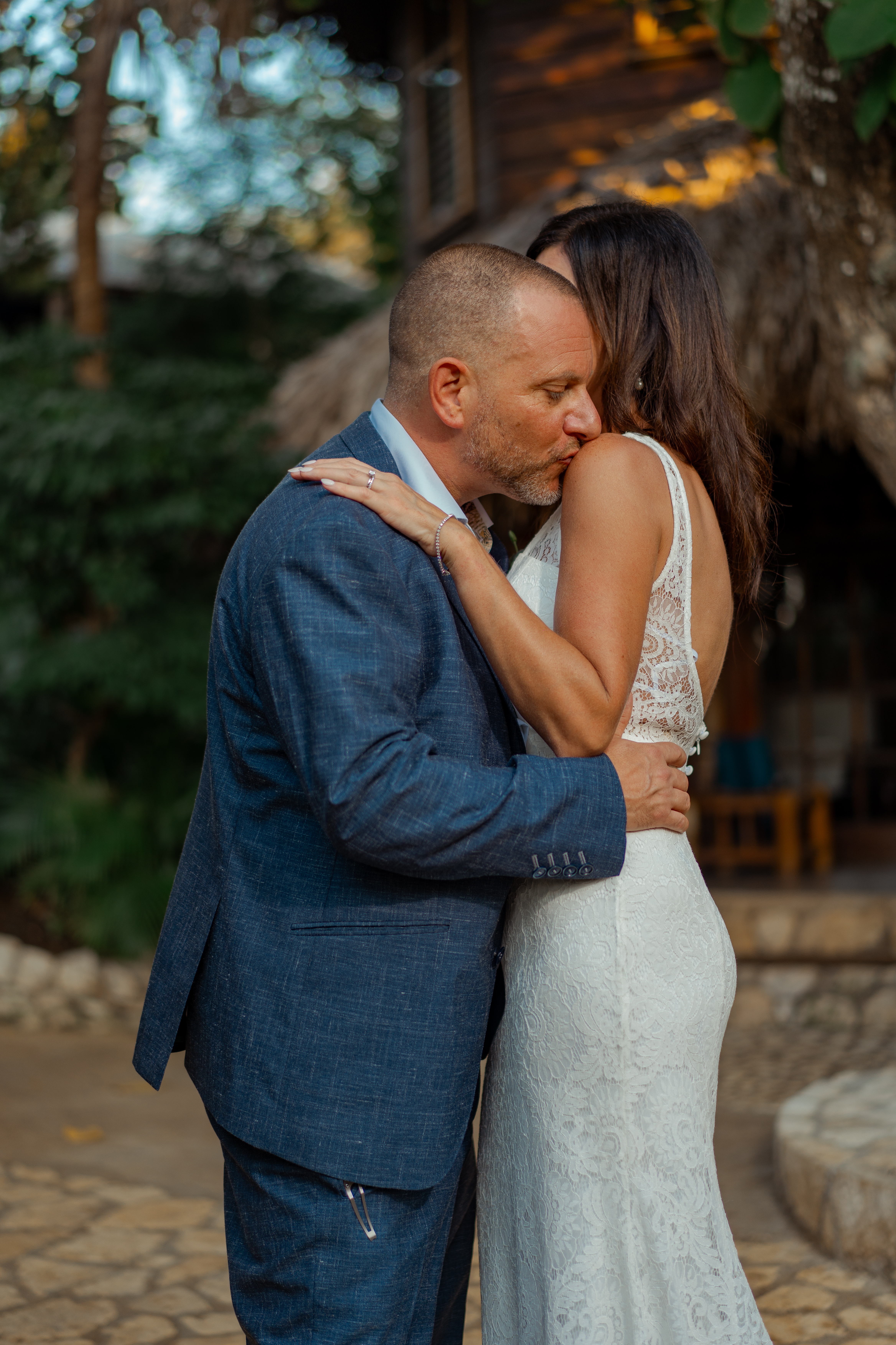 Joe kissing Sarah on the neck, thatched hut behind them